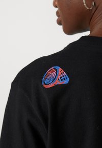 Black cotton t-shirt featuring a colorful graphic on the shoulder, depicting a rose within a geometric design with red and blue accents.