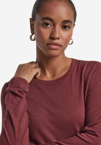 Maglione bordeaux lavorato a maglia con girocollo, maniche lunghe e polsini a coste. Presenta una texture liscia e una vestibilità aderente. Orecchini a cerchio dorati completano il look.