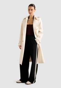 Donna che indossa un trench coat crema sopra una maglia marrone scuro, pantaloni neri a gamba larga con strisce beige ai lati e sneakers bianche, in piedi con una mano in tasca.