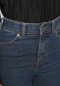 Jeans de denim azul oscuro de cintura alta con un diseño clásico de cinco bolsillos, que presentan herrajes en tono cobre y detalles de costura en contraste.