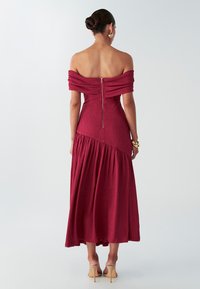 Bordeauxrotes Off-Shoulder-Kleid mit strukturierter Stoff, tailliertem Oberteil und gestuften Rock. Verfügt über einen Reißverschluss im Rücken und eine elegante Silhouette, kombiniert mit beigen Absätzen.