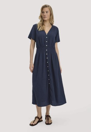 Frau trägt ein marineblaues Midi-Hemdkleid mit kurzen Ärmeln und geraffter Taille, kombiniert mit dunkelbraunen Sandalen.