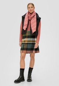 Zwarte gewatteerde vest over een roze langarmshirt, gecombineerd met een meerkleurige geruite rok en zwarte enkellaarsjes. Roze gestructureerde sjaal om de hals.