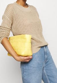 Béžový pletený svetr s kulatým výstřihem, spárovaný se žlutou tkátou kabelkou do ruky. Kabelka má zapletenou texturu a obdélníkový tvar.