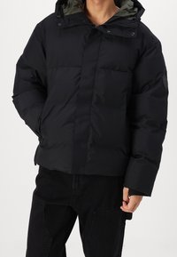 Mand iført en sort hætte-pufferjakke og sorte cargobukser, stående med den ene hånd i jakkelommen.