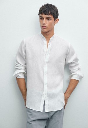 Giovane uomo con capelli scuri che indossa una camicia bianca con bottoni a maniche lunghe arrotolate e pantaloni grigi chiari, in piedi di fronte a uno sfondo semplice.