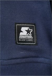 Sötétkék, kötött textúrájú anyag, amelyen egy fekete négyzet alakú logófelvarró található "STARTER BLACK LABEL" felirattal fehér színben és egy csillag ikonnal.