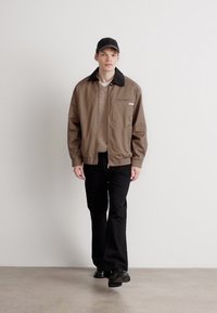 Rudas švarkas su juodu apykakle, su užtrauktuku, vilkimas virš šviesios megztinio ir juodų kelnių. Modelis stovi prieš neutralų foną.