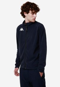 Giacca in pile blu navy con colletto alto, zip completa e logo bianco sul petto. Presenta maniche lunghe e una texture morbida.