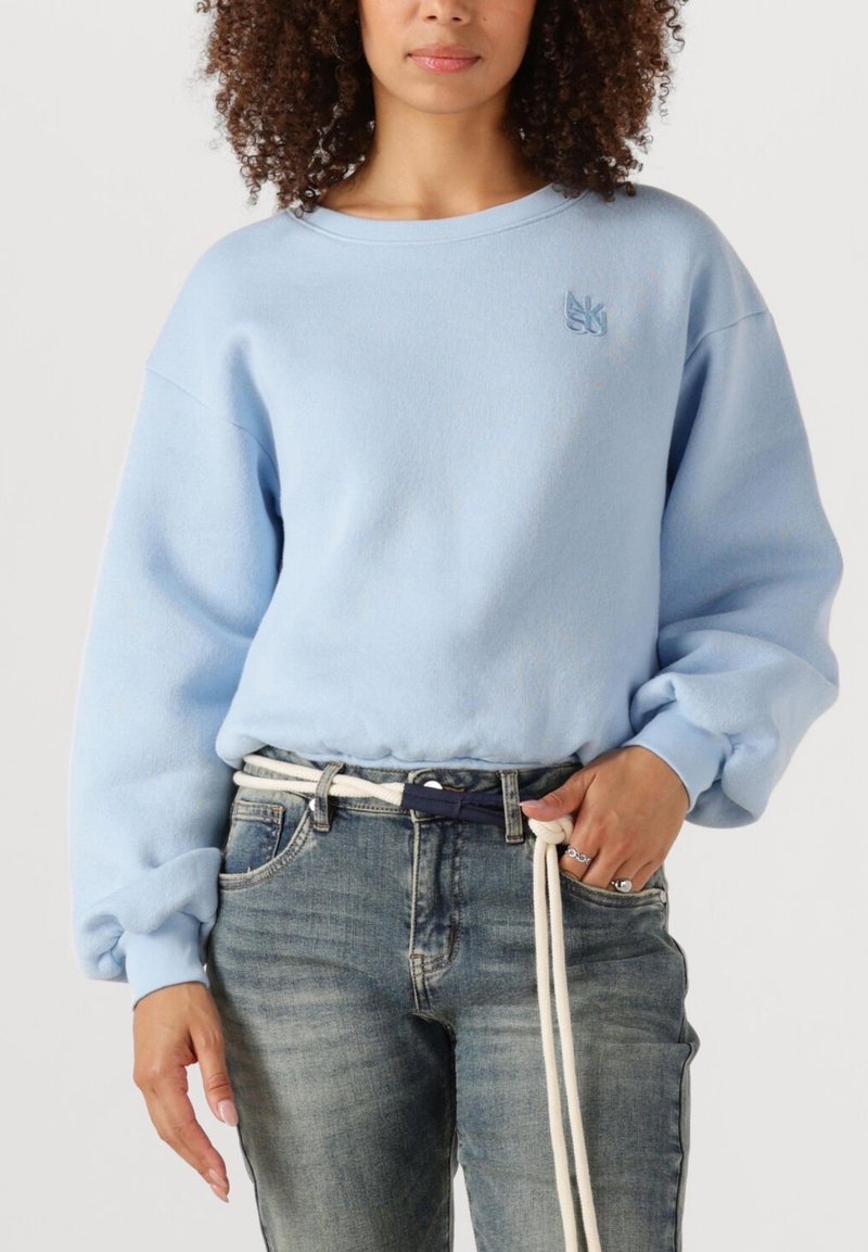 Frau in einem hellblauen, oversized Sweatshirt und hellblauen Jeans, die mit einem weißen Seilgürtel in der Taille verknotet sind.