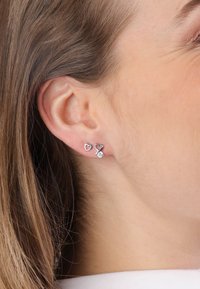 Zilveren oorbellen met hartvormige ontwerpen, voorzien van een rode accent en een centrale steen, gedragen aan de oorlel tegen een neutrale achtergrond.