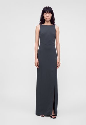Femme aux longs cheveux foncés portant une robe longue sans manches gris foncé avec une fente à l'avant et des chaussures noires ouvertes à l'avant, debout sur un fond clair.