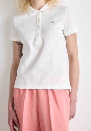Polo blanc en coton avec une patte de boutonnage à trois boutons et des manches courtes. Il présente un petit logo noir sur la poitrine gauche.