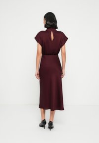 Robe midi bordeaux avec manches courtes, dos avec ouverture en trou de serrure, et taille cintrée. Texture lisse, ourlet évasé, associée à des talons noirs.
