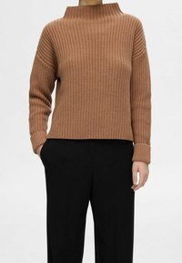 Pull en tricot marron avec un col montant, une texture côtelée et des épaules tombantes. Associé à un pantalon noir à jambes larges.