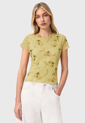 Nuori vaaleatukkainen nainen pukeutuu vaaleanvihreään kukkakuvioiseen verkkotoppiin ja valkoisiin korkeavyötäröisiin housuihin, käsi taskussa, harmaalla taustalla.