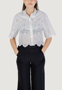 Maglietta bianca corta con chiusura a bottoni, caratterizzata da design floreali tagliati al laser e maniche ad arco. Abbinata a pantaloni neri a vita alta.