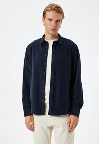 Camisa de pana azul marino con mangas largas, cierre de botones y un único bolsillo en el pecho, usada sobre una camiseta de cuello redondo de color claro.