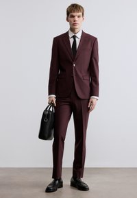 Μπορντό κοστούμι με εφαρμοστή γραμμή, κλείσιμο με ένα κουμπί και πέτο με εγκοπή. Μαύρη δερμάτινη τσάντα και παπούτσια ολοκληρώνουν την εμφάνιση. Λευκό πουκάμισο και μαύρη γραβάτα.