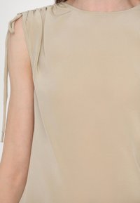 Top beige senza maniche con dettagli arricciati sulle spalle e lacci, realizzato in un tessuto liscio e lucido che riflette la luce in modo sottile.