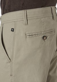 Beige Baumwollhose mit strukturiertem Gewebe, ausgestattet mit einer Gesäßtasche mit Knopf und kontrastierenden Nähten. Kleines Logotag sichtbar.