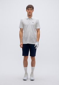 Λευκό πόλο πουκάμισο με υφασματικό σχέδιο και μαύρο λογότυπο, συνδυασμένο με σκουρόχρωμο σορτς. Το μοντέλο φοράει λευκά παπούτσια και γάντι, στέκεται όρθιο.