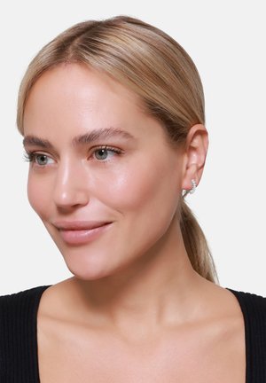 Øredobber med en buet design, prydet med små klare edelstener i sølvfarget metall, bæres av en lys hudet person.
