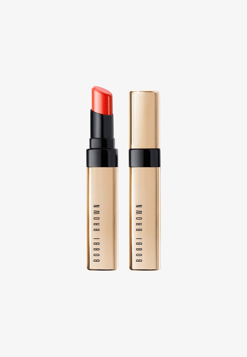 Lippenstift in einer goldenen Hülse mit schwarzem Boden. Die Lippenfarbe ist leuchtendes Korall mit satinierter Oberfläche. Die Verpackung hat ein minimalistisches Logo.