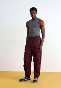 Haut gris sans manches, pantalon bordeaux ample avec poches latérales, baskets noires avec accents blancs. Matière lisse, coupe décontractée, design minimal.