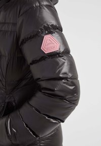 Manga de una chaqueta acolchada negra y brillante con un parche en forma de pentágono rosa etiquetado como "JACK IT" en la parte superior del brazo, sobre un fondo liso.