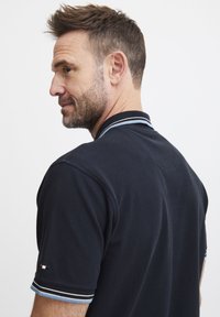 Marineblå polo med teksturert stoff, som har en klassisk krage og kontrasterende blå og hvite striper på ermene, med et lite logo.