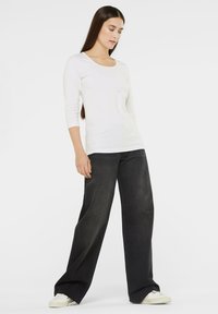 Weißes Langarmoberteil mit Rundhals und kombiniert mit weit geschnittenen, dunklen Jeans sowie weißen Sneakern, was einen einfachen, lässigen Stil zeigt.