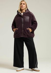 Fioletowa polarowa kurtka z kapturem, z gładką wstawką na piersi i dwoma przednimi kieszeniami. Noszona z czarnymi spodniami o szerokich nogawkach i białymi trampkami.
