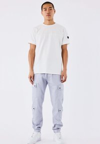 Giovane uomo in piedi, indossa una maglietta bianca a maniche corte semplice, pantaloni cargo grigi chiari con tasche laterali e sneakers bianche su uno sfondo bianco.