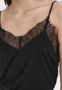 Top nero senza maniche con scollatura decorata da pizzo. Il pizzo presenta motivi floreali e bordi sfrangiati, con spalline sottili e regolabili. Tessuto morbido.