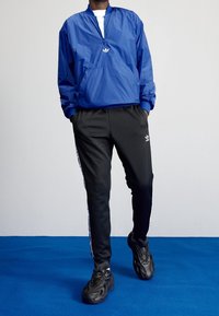 Modrý větruodolný kabát se zipem a vyšitým logem; doplněný černými tepláky s bočními pruhy a černými sportovními botami.