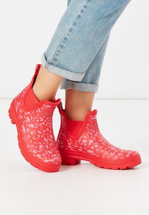 Nohy v červených kotníkových gumácích s bílým puntíkovaným vzorem a vyhrnutých světle modrých džínách na bílém podlaze.