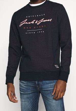 Czarna bluza z napisem „Jack & Jones Originals DNM Goods Since 1990”, noszona z niebieskimi dżinsami, pokazująca górną część ciała i ręce.