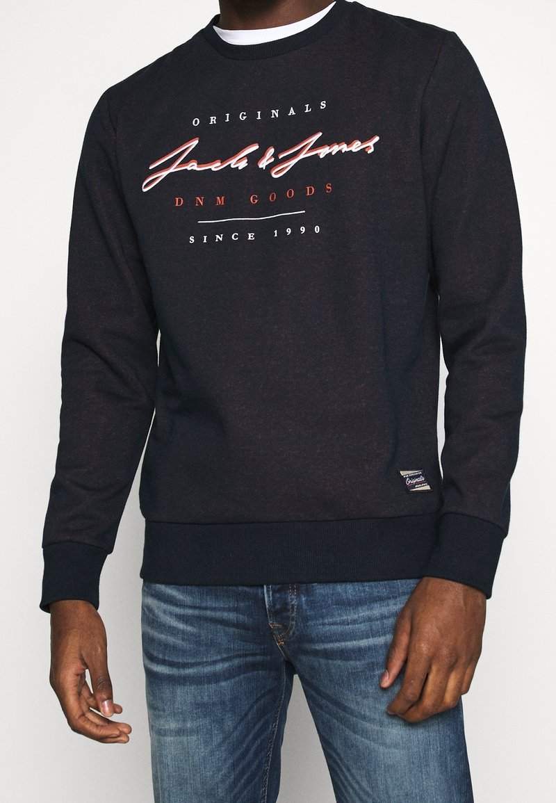 Sweat noir avec le texte « Jack & Jones Originals DNM Goods Since 1990 », porté avec un jean bleu, montrant le haut du corps et les mains.