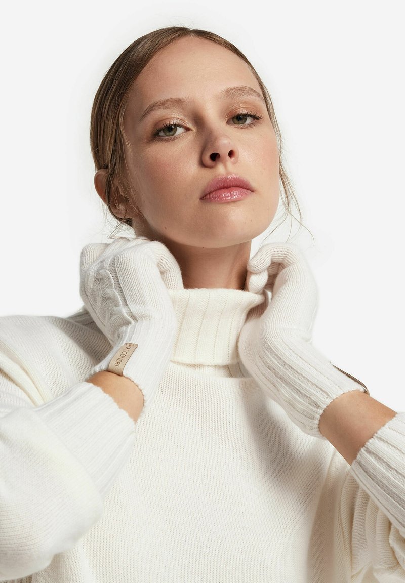 Białe wełniane rękawiczki bez palców o prążkowanej strukturze z naszywką logo, noszone z dzianinowym swetrem z wysokim kołnierzem w miękkim białym kolorze.