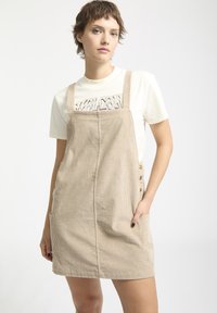 Beige corduroy jurk met schouderbanden, voorzakken en knoopaccenten. De textuur is geribbeld en het ontwerp is getailleerd.