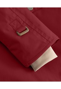 Manga de chaqueta de lluvia roja con un puño interno beige doblado, que presenta una hebilla de metal y detalles cosidos en la zona de la muñeca. Textura suave.