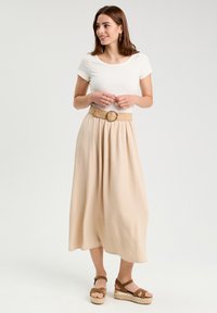 Jupe beige fluide en tissu texturé, taille cintrée avec une boucle de ceinture ronde, associée à un haut blanc ajusté et des sandales compensées.