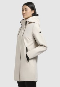 Veste imperméable beige clair avec capuche, accents noirs, deux poches zippées et design épuré. Présente un éclat subtil et une texture lisse.