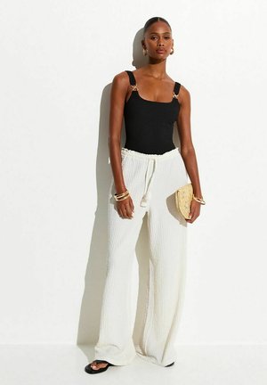 Mujer con top negro sin mangas, pantalones anchos blancos con textura, sandalias negras, sosteniendo un pequeño clutch beige sobre fondo blanco.