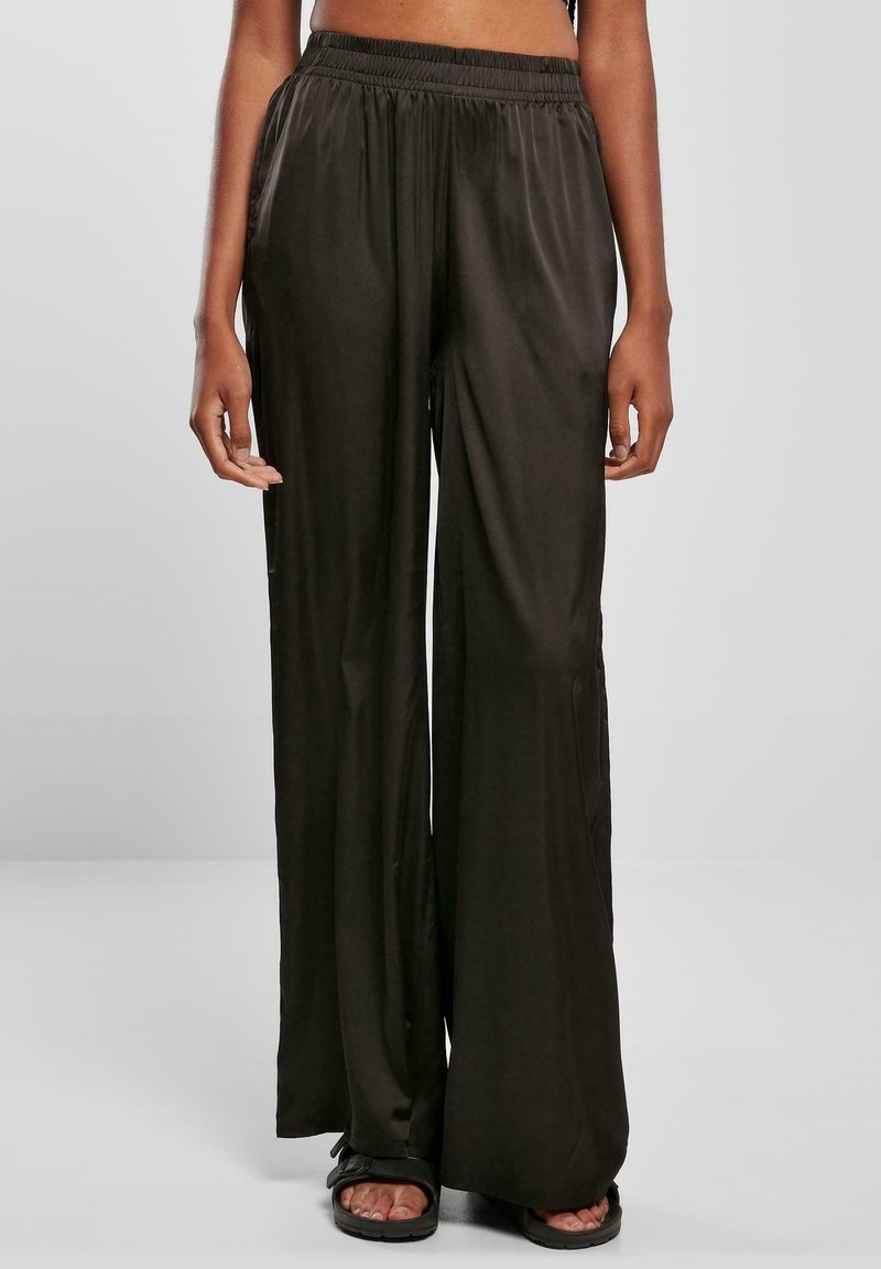 Personne portant un pantalon large en satin noir ample avec une taille haute élastique et des sandales noires, debout devant un fond gris.
