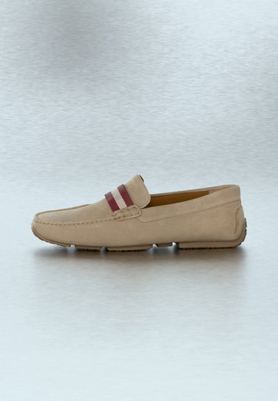 Mocassin en daim beige avec des détails cousus et une sangle en tissu rayée rouge et blanche sur le dessus, posé sur une surface argentée texturée.