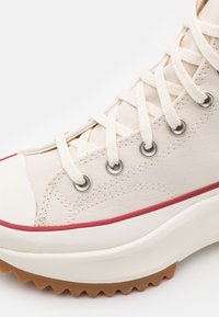 Plátěné vysoké tenisky v off-white, s béžovými tkaničkami, stříbrnými očky a červenými akcenty podél gumové podrážky a špičky.