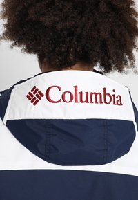 Personne aux cheveux bouclés portant une veste bleu marine et blanche avec un logo Columbia rouge brodé sur le col arrière.