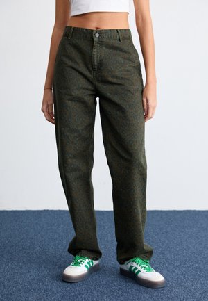 Woman wearing high-waisted dark green leopard-print pants with white sneakers featuring green stripes and laces, standing on blue carpet.